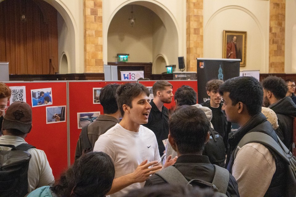 Students talking with visitors at a busy society showcase.