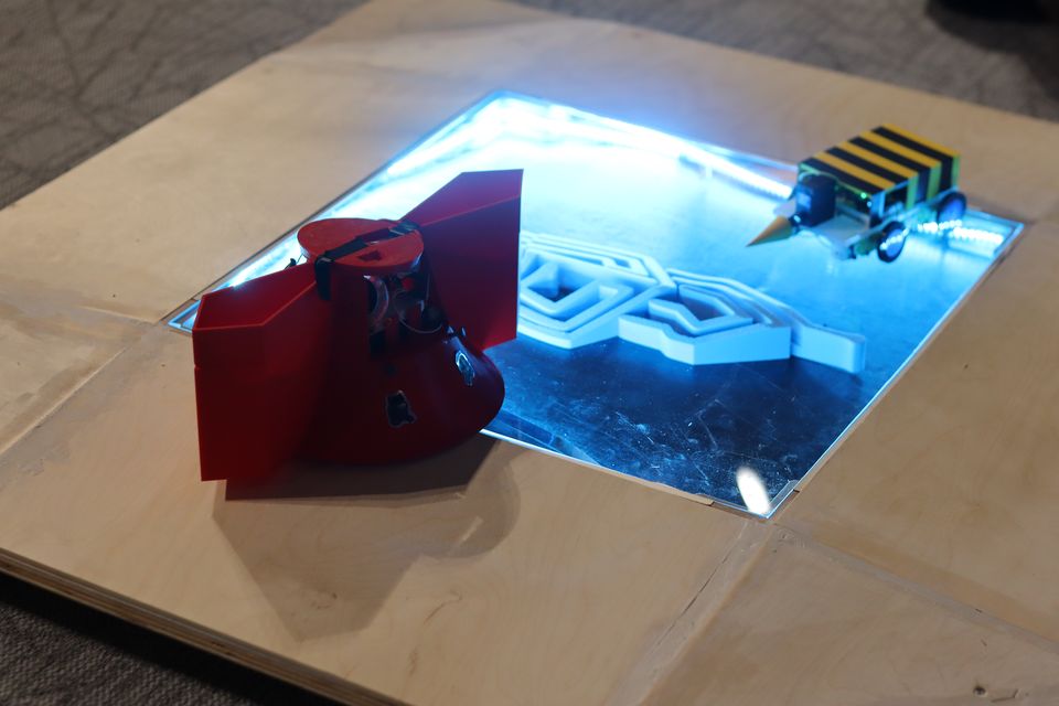 A red student-built robot positioned at the edge of an illuminated arena.