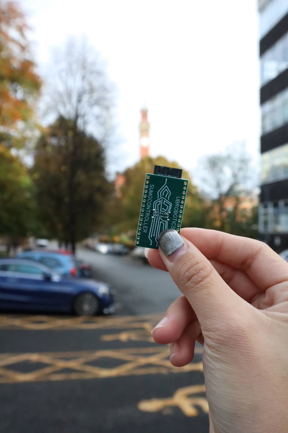 A hand holding a UBRobotics printed circuit board outdoors on campus.