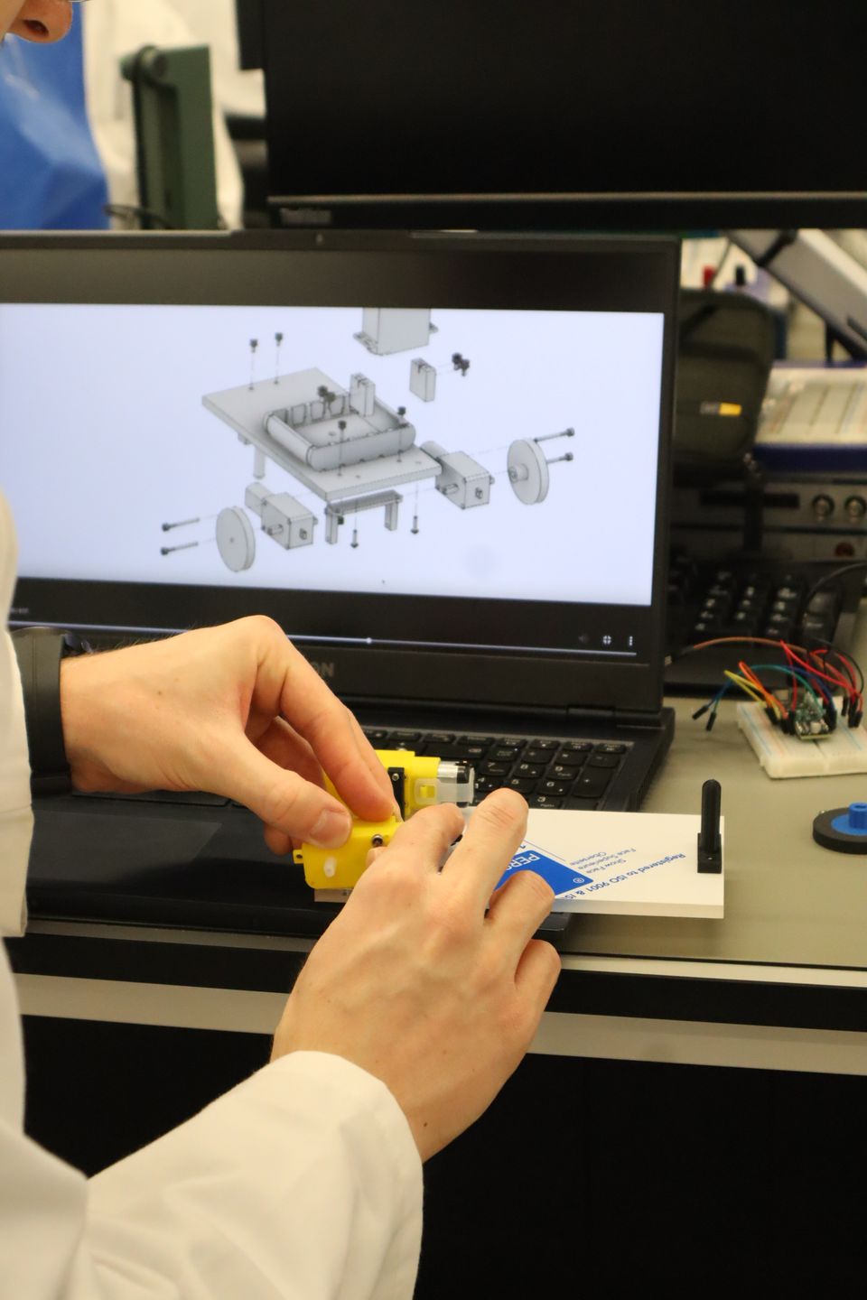 Hands assembling yellow mechanical parts beside a laptop.